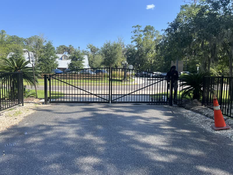 Double aluminum driveway gate across an asphalt drive framed by palms and live oaks