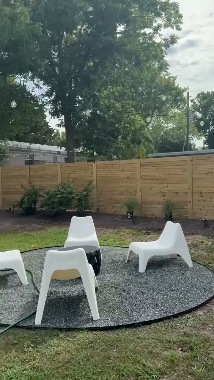 Horizontal wood fence wrapping a backyard with a round gravel patio, white chairs, and string lights overhead