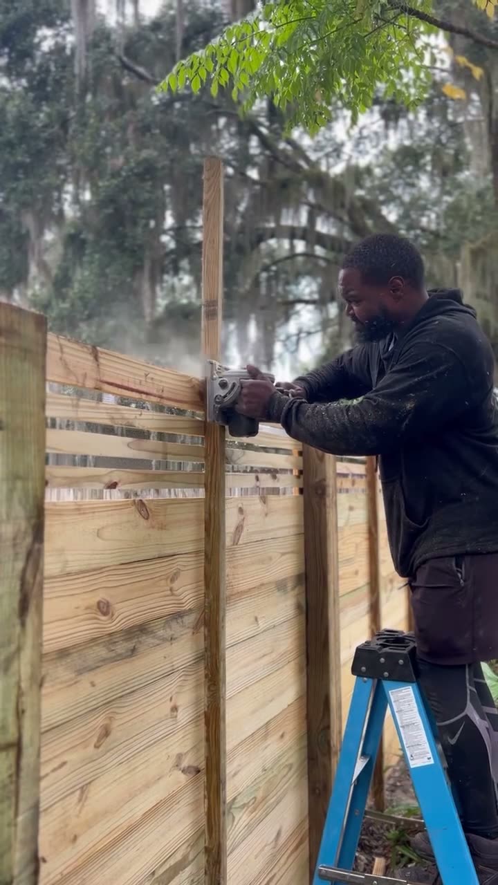 Dynamic Fence crew member on a ladder cutting a fence board with a circular saw, sawdust in the air