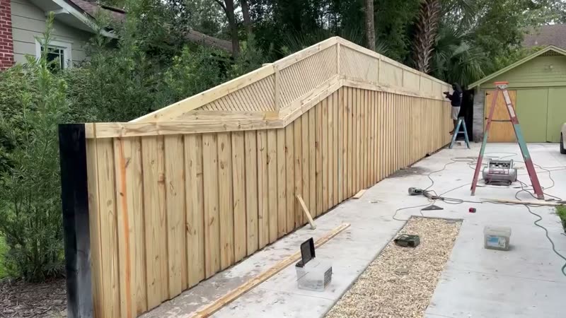 Tall wooden privacy fence with an angled lattice top section stepping up along a concrete driveway, black end post in the foreground